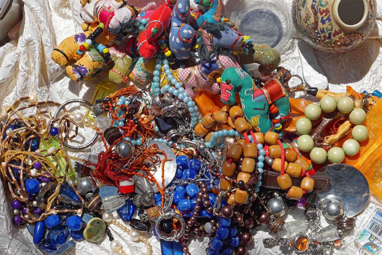 A vibrant collection of handmade jewelry and trinkets displayed on a table at a flea market. The assortment includes colorful beaded necklaces, bracelets with wooden and pearl beads, fabric dolls, and various other small decorative items. The background features a patterned cloth, enhancing the rich and eclectic mix of textures and colors. The scene captures the lively and artistic spirit of the market.